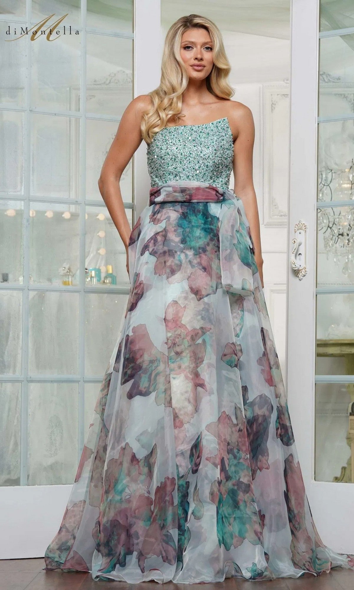 A woman stands indoors before glass doors, wearing the Rina Di Montella RD3213 long formal dress with a strapless beaded bodice and flowing floral-printed organza skirt.